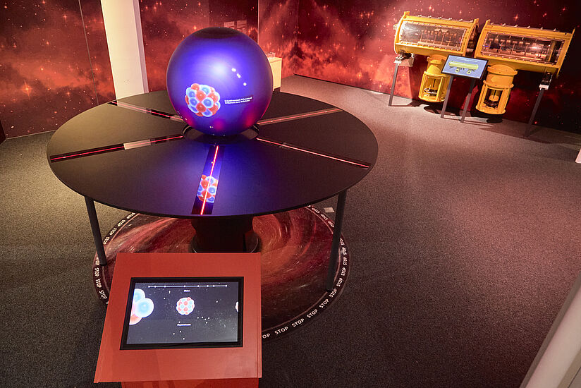 Ein interaktives Exponat im Teilchenzoo des Deutschen Museums Bonn, bestehend aus einer Kugel auf einem runden Tisch und einem Bildschirm.