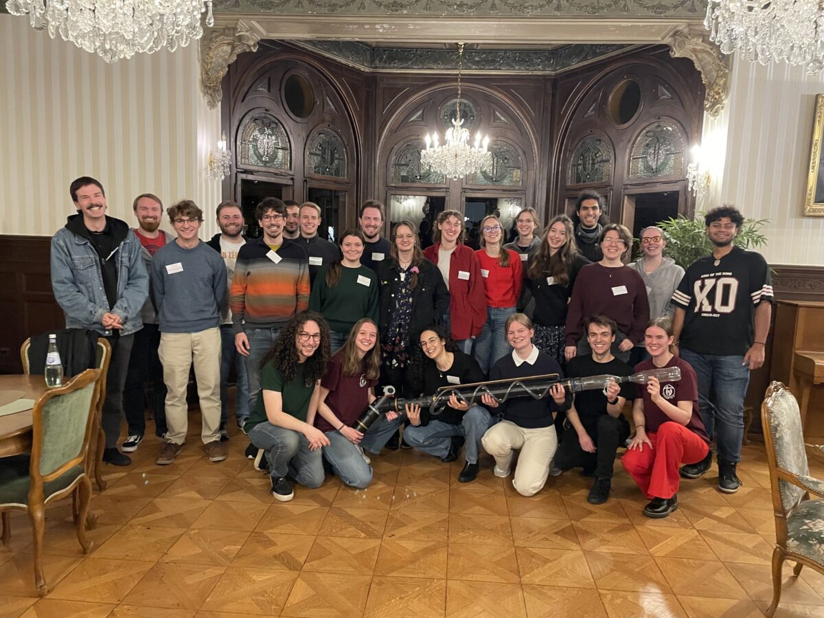 Gruppe junger Menschen posiert lächelnd in elegantem Saal, einige halten ein langes technisches Objekt.